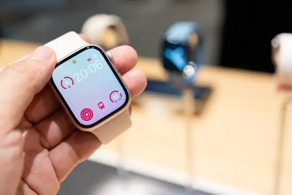 Une main tient une montre connectée affichant l'heure 20:06, des indicateurs d'activité et le logo watchOS 26.